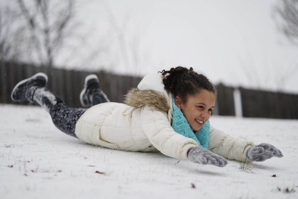 Girl sliding down the snow on her stomach in white jacket and green scarf.